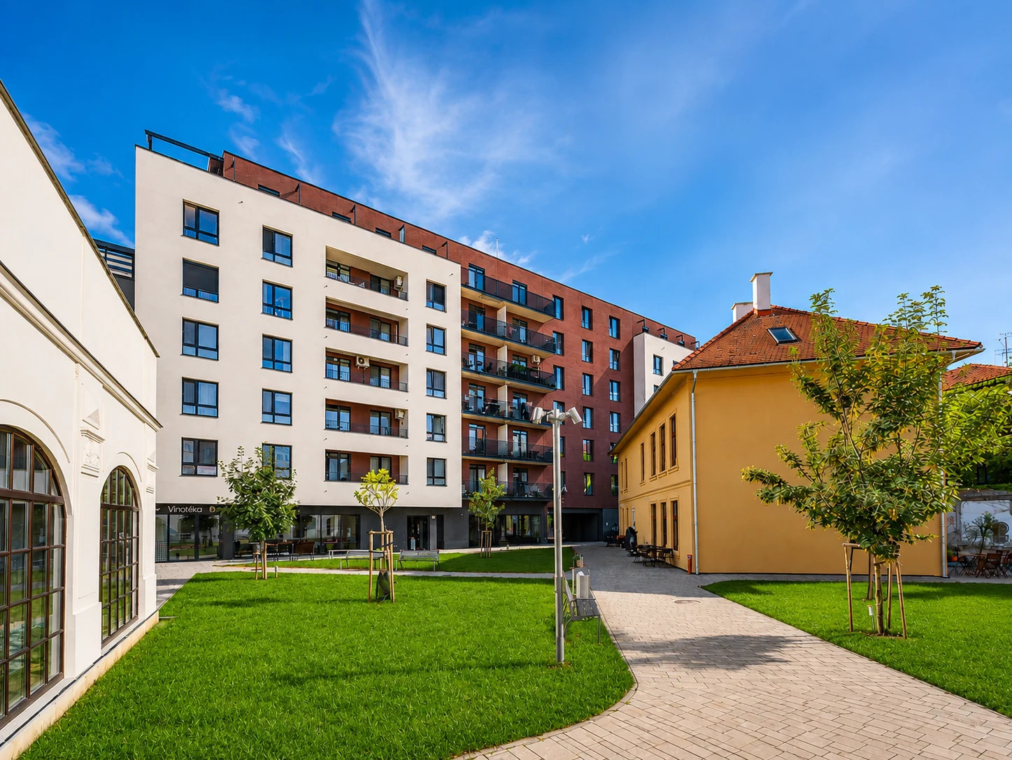 Moderný viacposchodový bytový dom s balkónmi, vnútorným nádvorím a ľuďmi prechádzajúcimi za súmraku.