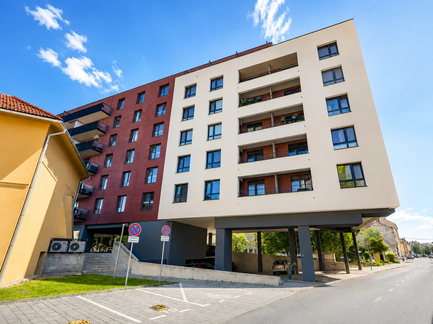 Moderná budova s červenou strechou, ľudia prechádzajú po chodníku v parku pod jasnou oblohou.