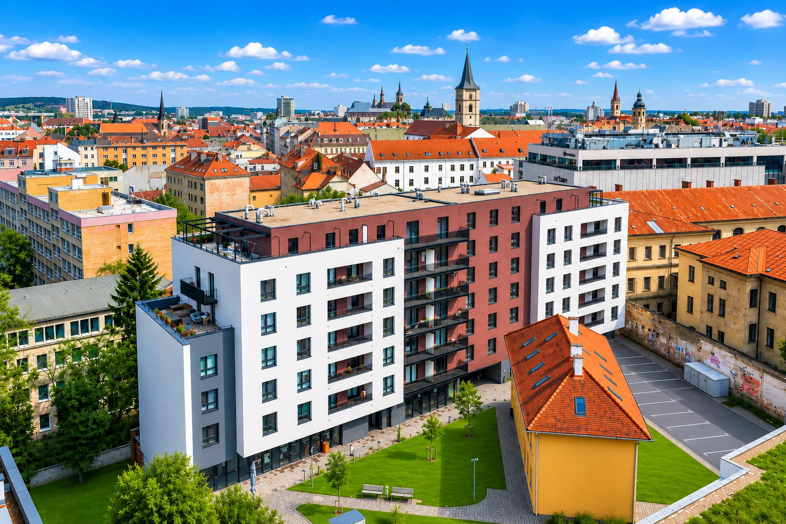 Panoramatický pohľad na mesto s bytovým domom, červenými strechami a vežami pod modrou oblohou.